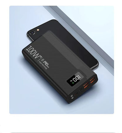 Black textured 100W power bank with an LED display showing 100% charge, resting on a black smartphone.